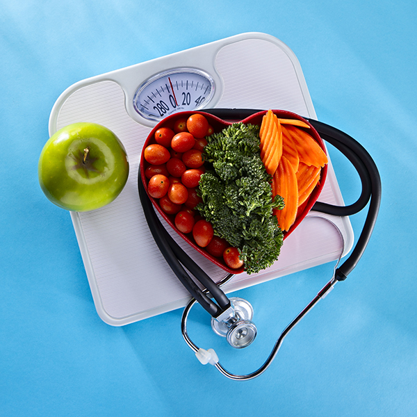 This is an overhead photograph of vegetables in a heart bowl sitting next to dumbells on a scale with a blue background. This image symbolizes healthy living and eating lifestyle